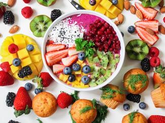 A bowl with strawberries, blueberries, kiwi and surrounding health food such as muffins, pineapples, and blackberries